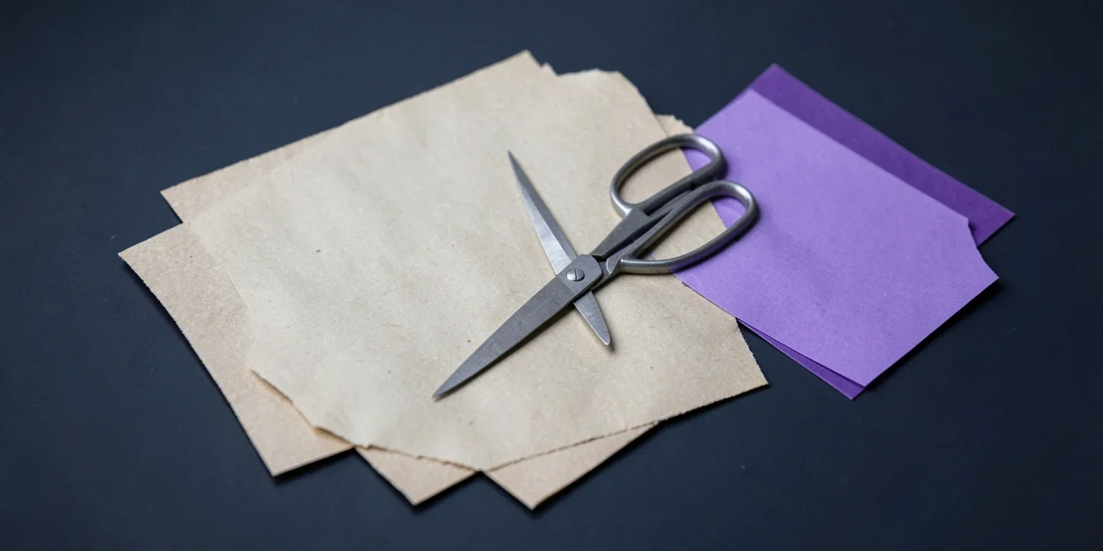Dos hojas de papel crudo casi idénticas superpuestas, una hoja violeta aparte y unas tijeras abiertas encima Dos hojas de papel crudo casi idénticas superpuestas, una hoja violeta aparte y unas tijeras abiertas encima