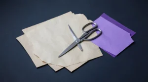 Dos hojas de papel crudo casi idénticas superpuestas, una hoja violeta aparte y unas tijeras abiertas encima