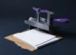 Prensa de tornillo metálica con manivela violeta sobre una pila de hojas de papel blanco